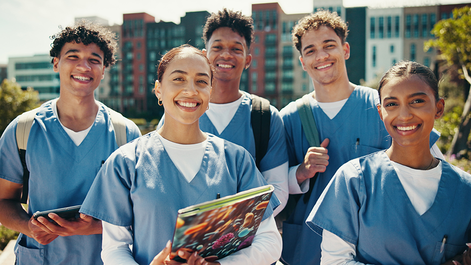 Group of diverse Licensed Vocational Nursing (LVN) students in blue scrubs smiling outdoors on campus with city buildings in the background